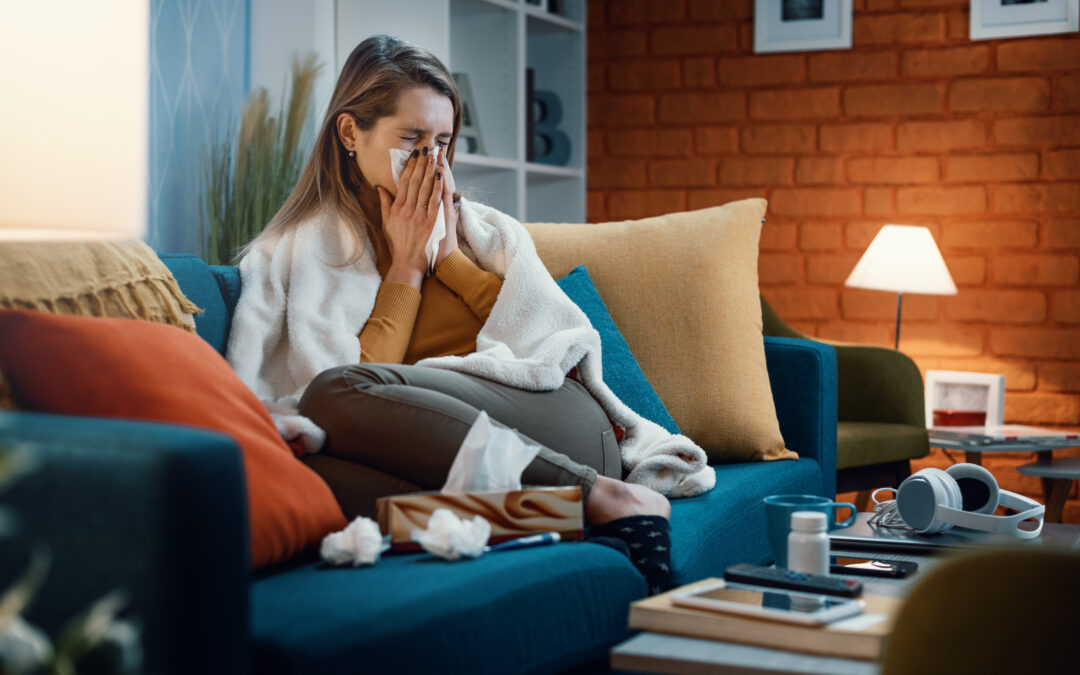 Woman sitting on couch, blowing nose or sneezing, surrounded by tissue, allergy medicine, and covered in blanket or shawl.